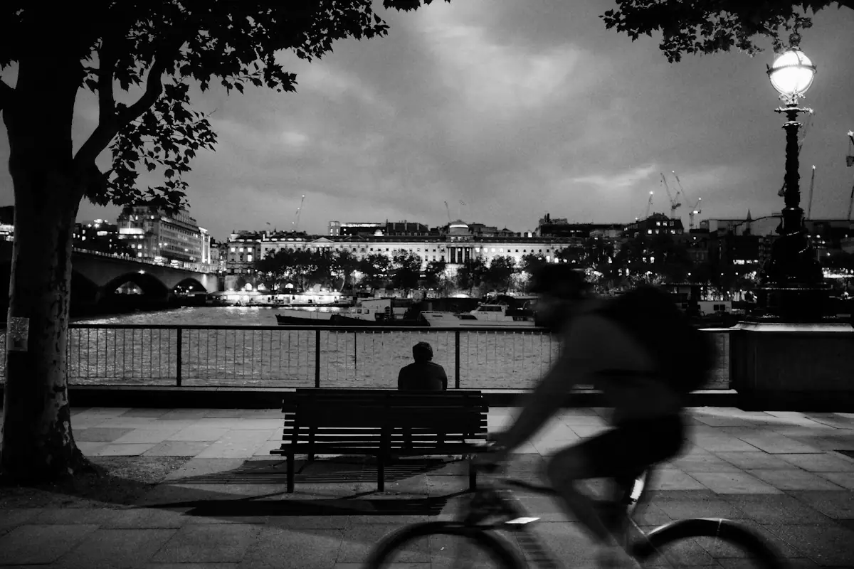 A person resting on a riverside bench at night while a cyclist passes.